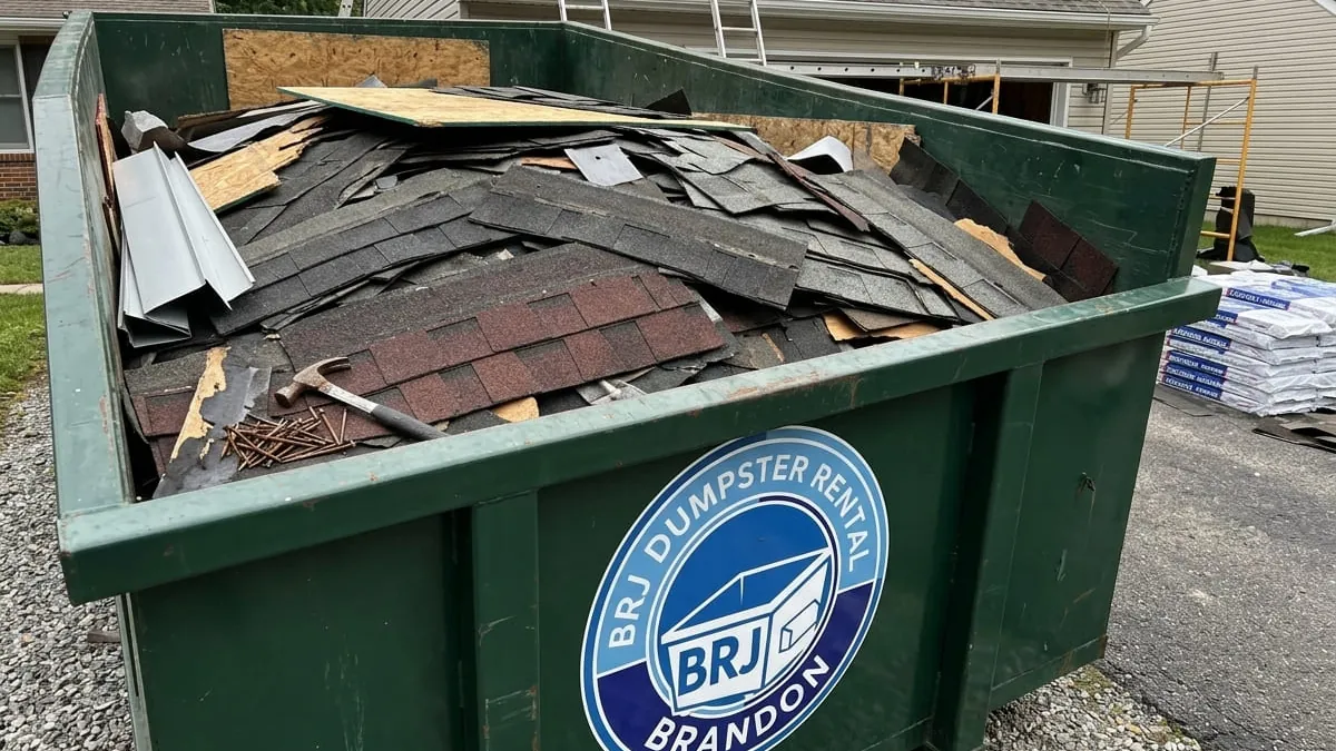 Roofing dumpster rental in Brandon: a 15 yard roll-off set on plywood tight to the eave of a 1970s ranch off Parsons Avenue, ground tarp laid out and torn-off shingles loaded flat and level below the fill line. Roofing dumpster rental in Brandon: a 15 yard roll-off set on plywood tight to the eave of a 1970s ranch off Parsons Avenue, ground tarp laid out and torn-off shingles loaded flat and level below the fill line.
